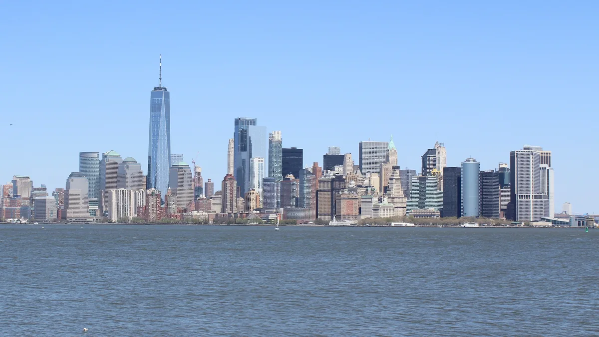 Lower Manhattan skyline with One World Trade Center above New York Harbor under a clear blue sky.